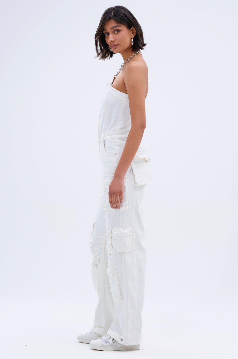 A woman with a short dark bob stands sideways against a white background, wearing the LINDSAY DENIM CARGO JUMPSUIT IN WHITE with multiple pockets, white sneakers, and a necklace for a stylish utility look.