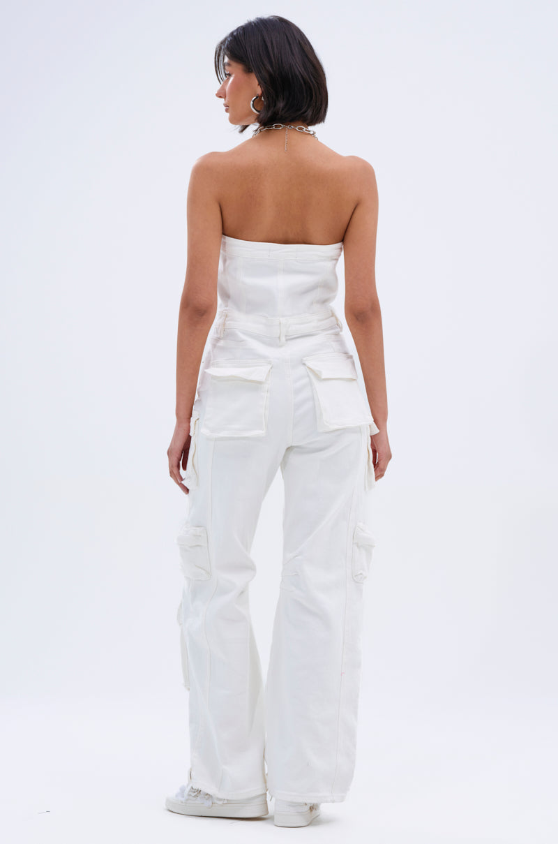 A woman with short dark hair stands facing away, wearing the LINDSAY DENIM CARGO JUMPSUIT IN WHITE— a strapless, pocket-detailed piece— paired with white sneakers against a plain light background, showcasing modern utility style.