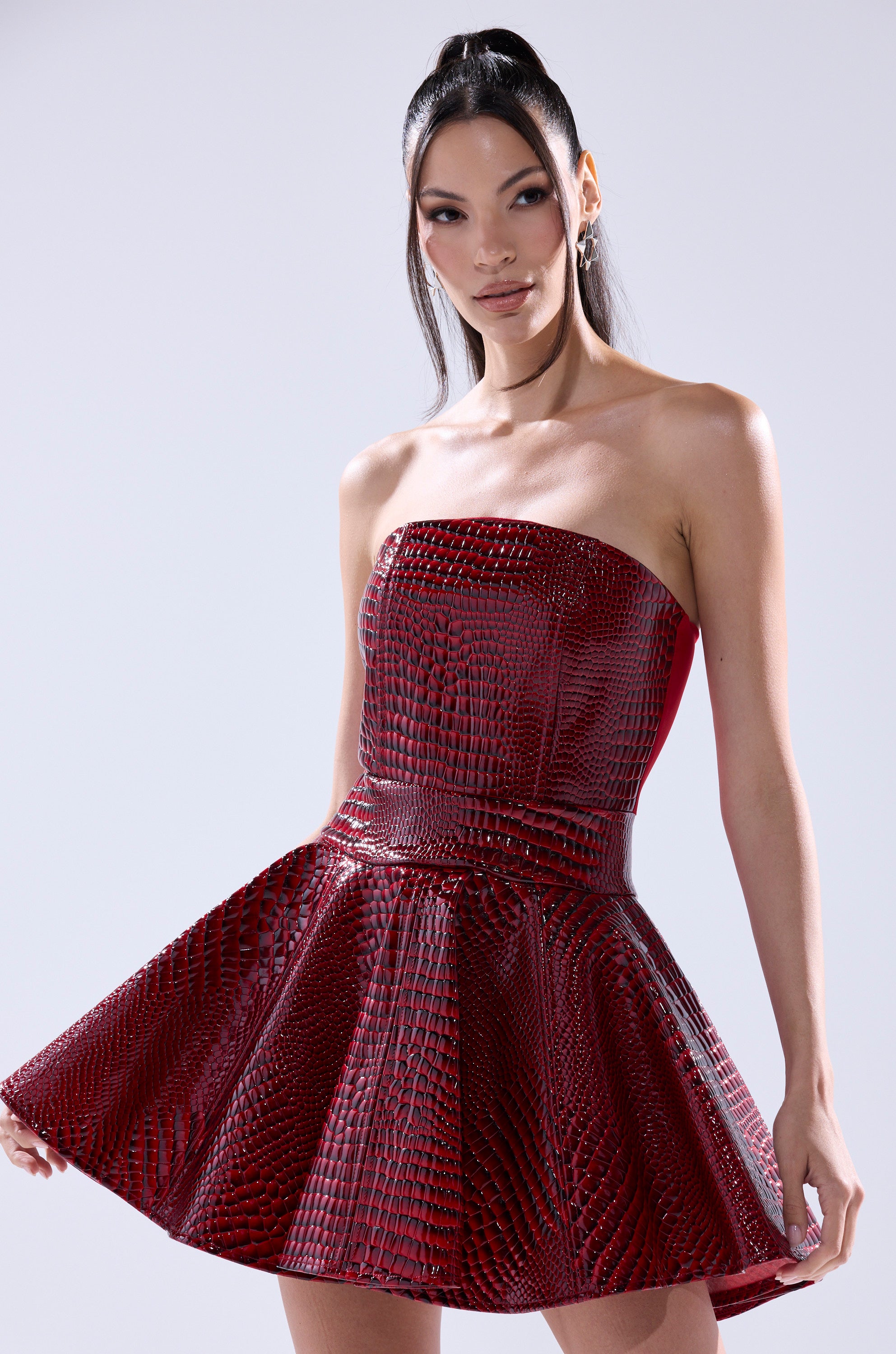 A woman wears the VAL PU MINI DRESS IN CROC PRINT by AKIRA Label, a strapless red faux crocodile leather dress, standing against a light background, holding the skirt with one hand and smiling slightly.