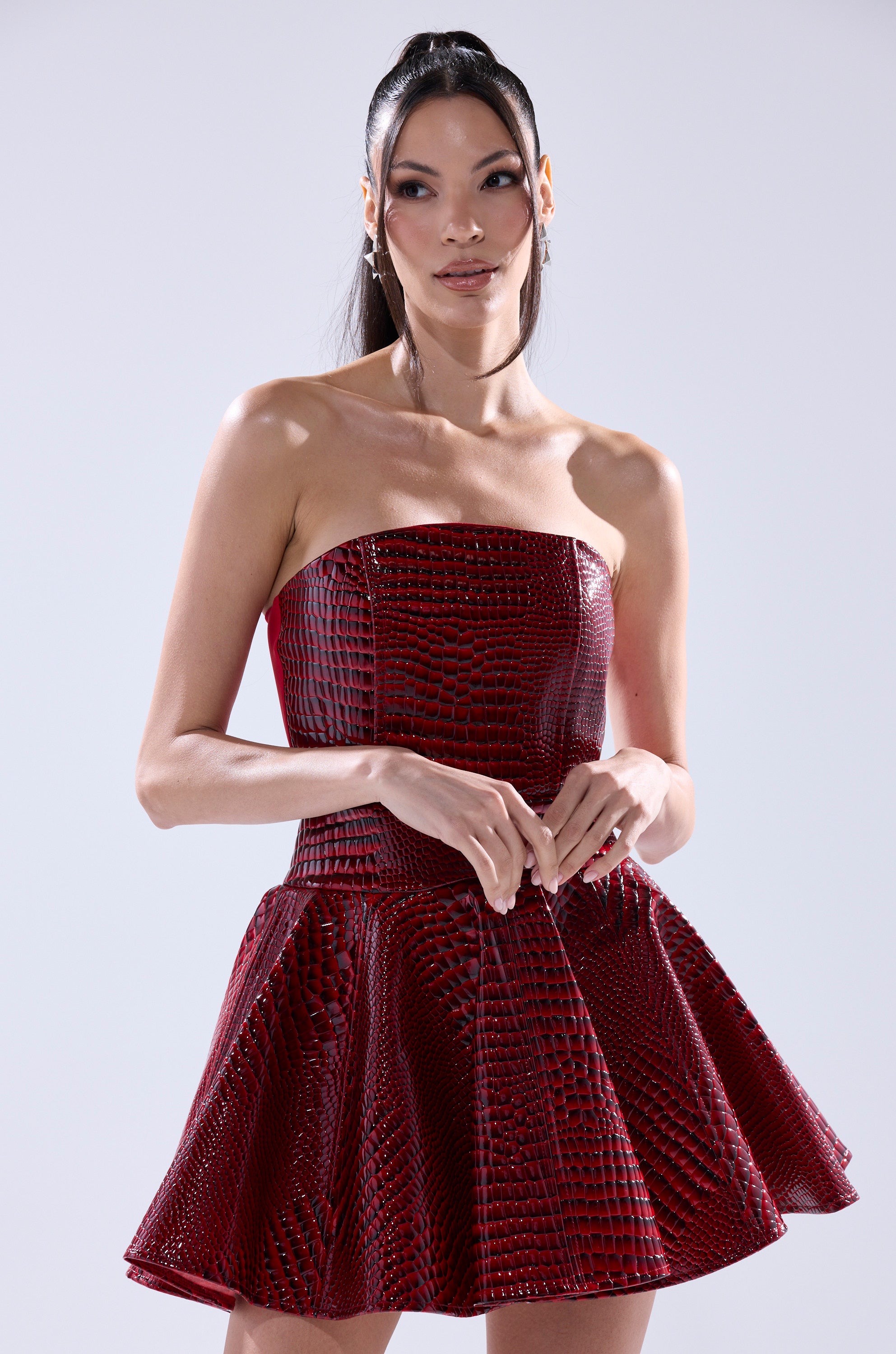 A woman with long dark hair in a high ponytail wears the VAL PU MINI DRESS IN CROC PRINT, featuring a strapless corset-style bodice and flared skirt. She stands against a plain light background, looking slightly to the side and smiling.