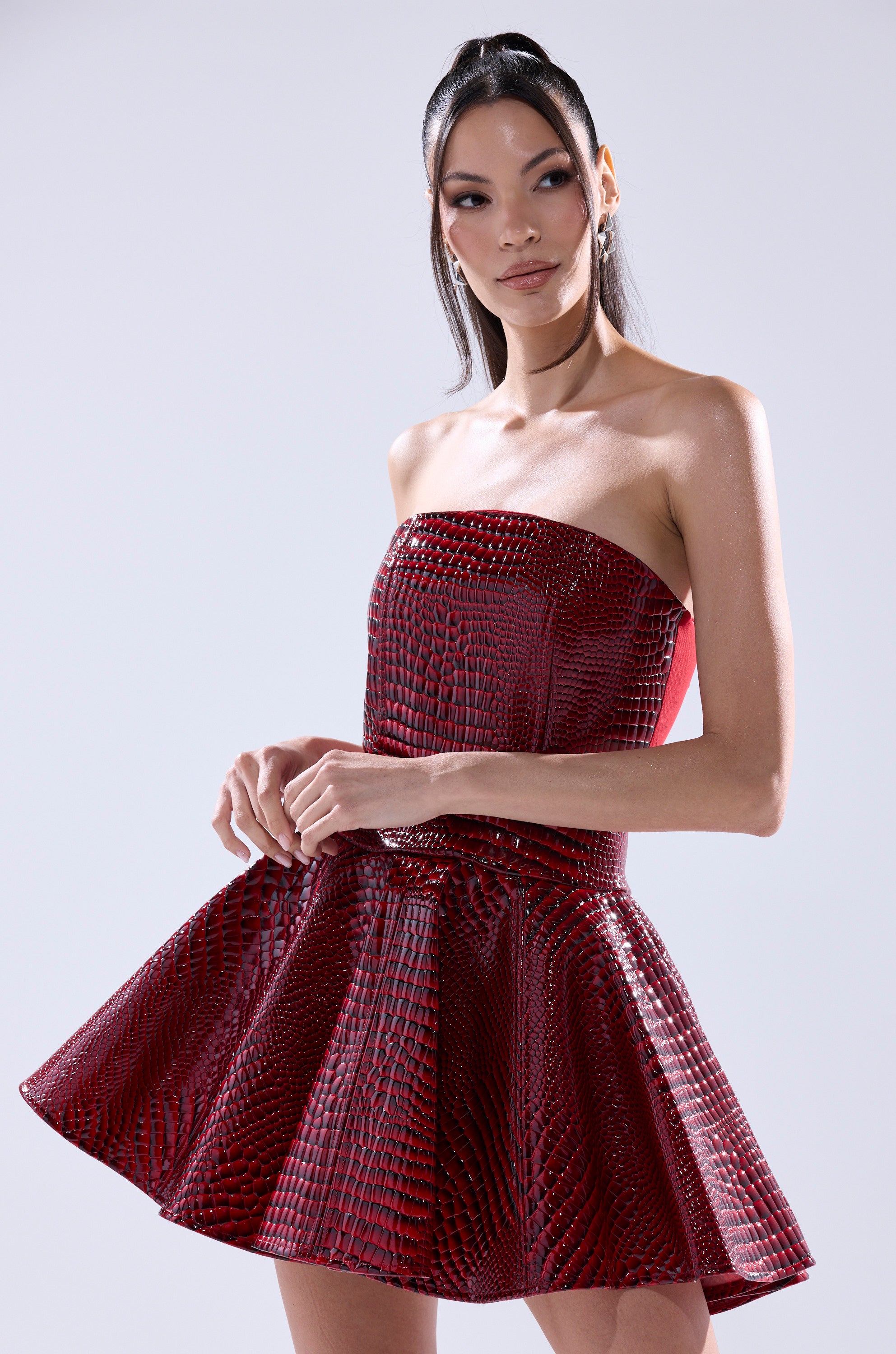 A woman poses against a light background in the VAL PU MINI DRESS IN CROC PRINT—a strapless red mini dress with a corset-style bodice, flared skirt, and textured faux croc finish—looking to the side with hands gently clasped.