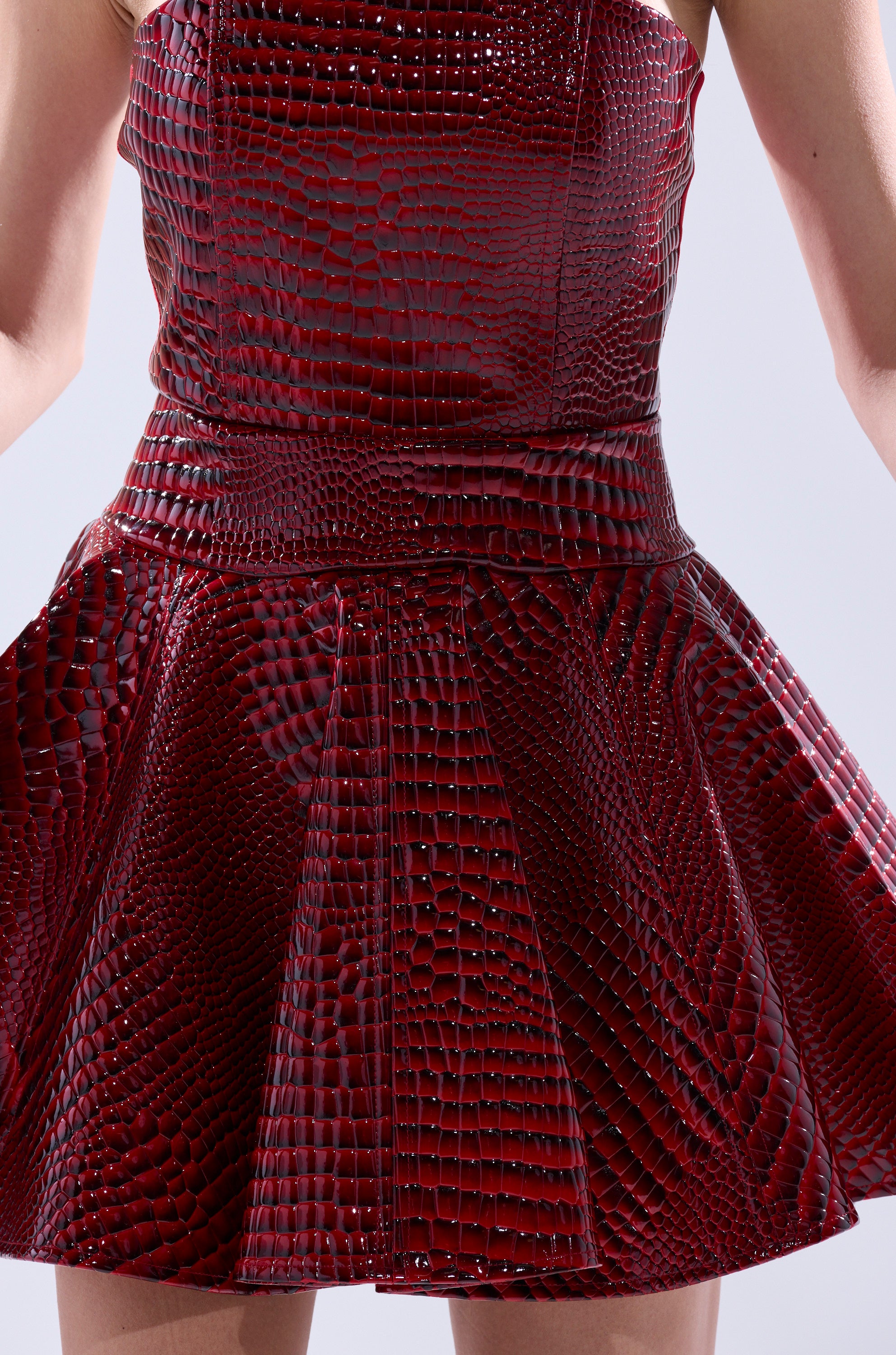 A person is shown from the waist down wearing the VAL PU MINI DRESS IN CROC PRINT, a shiny, textured dark red faux crocodile leather mini dress with a flared skirt, set against a plain background.