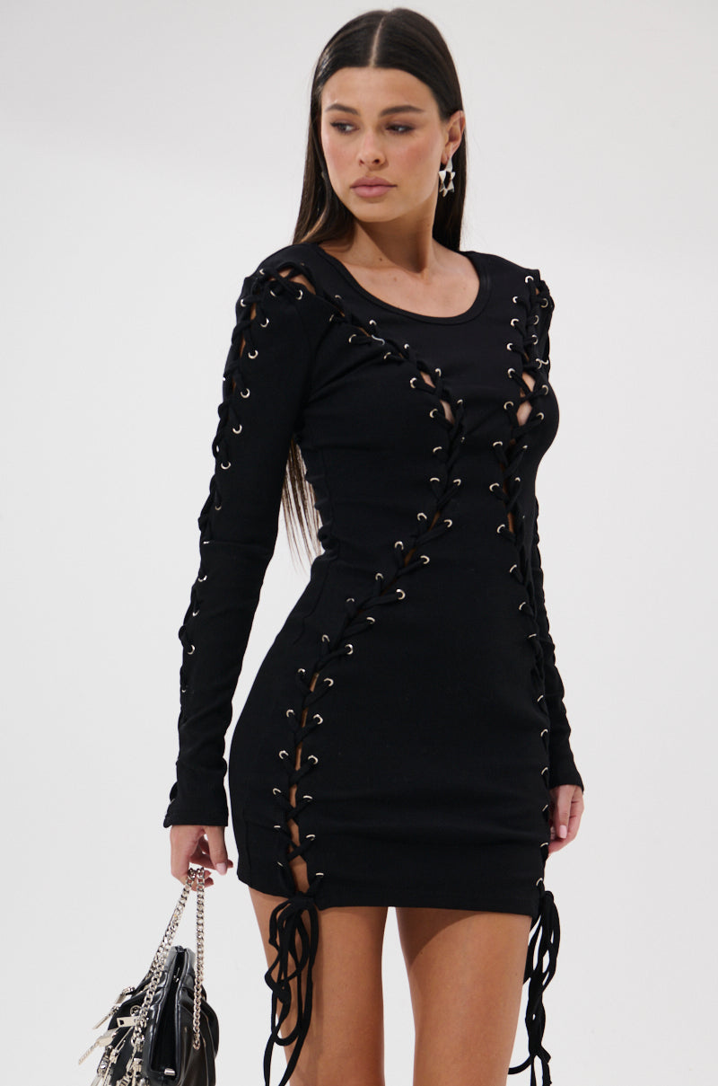 A woman stands against a plain white background, wearing the RUN THIS TOWN LACE UP MINI DRESS with long sleeves and a lace-up front, holding a black handbag and looking to the side.