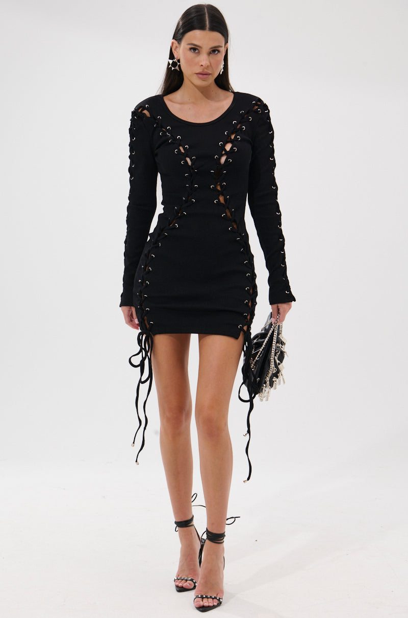 A woman stands against a white background wearing the RUN THIS TOWN LACE UP MINI DRESS, styled with long sleeves, a fitted silhouette, black high-heeled sandals, and carrying a small handbag.