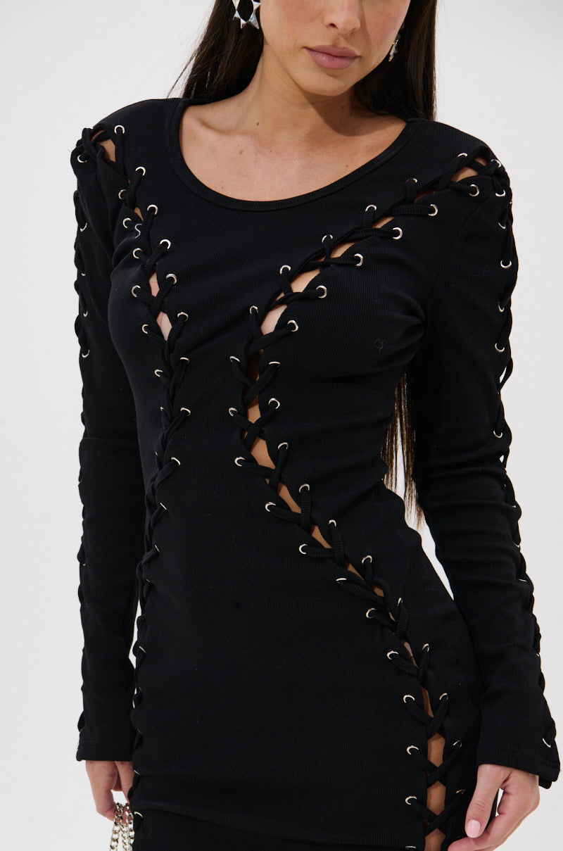A woman wears the RUN THIS TOWN LACE UP MINI DRESS, a fitted black bodycon mini dress with long sleeves, silver grommets, and crisscross lace-up diagonal cutouts on the front and sleeves. The background is plain white.