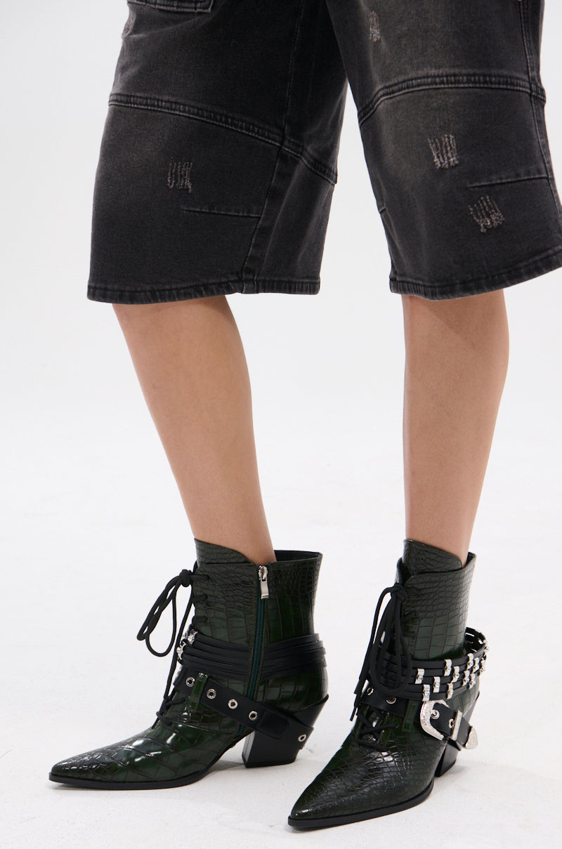 A person wearing black distressed knee-length shorts and AZALEA WANG JAVELIN GREEN WESTERN BOOTIE with buckles, laces, and silver hardware stands on a white background.