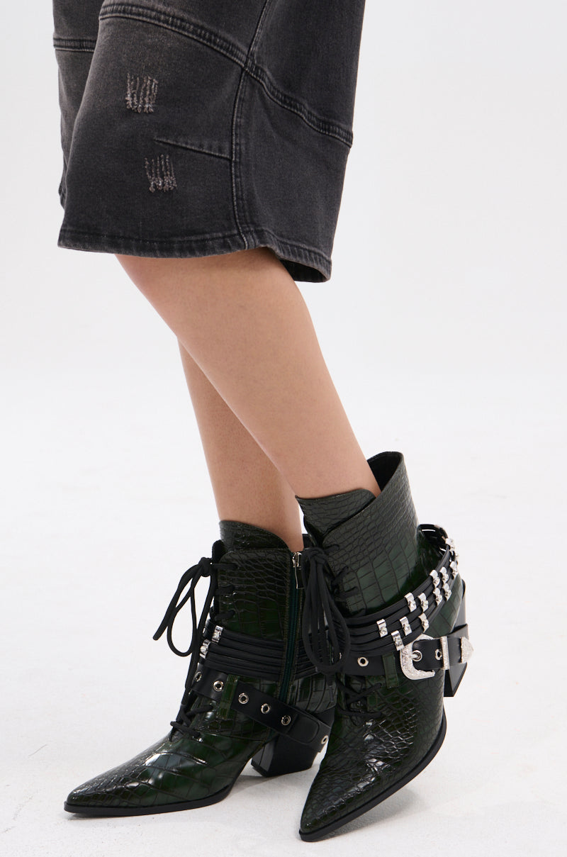 A person wears AZALEA WANG JAVELIN GREEN WESTERN BOOTIE with pointed toes and buckle details, styled with dark distressed knee-length shorts and socks, standing against a plain white background.