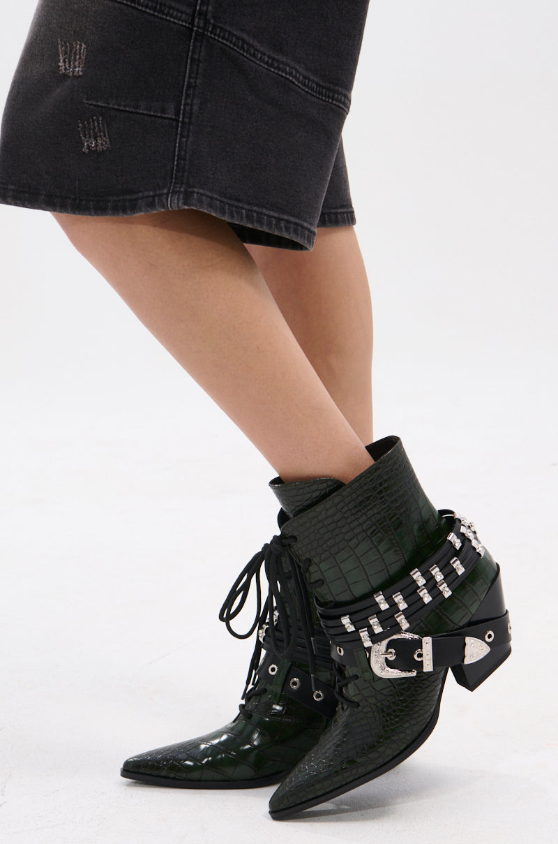 A person in a knee-length, faded black denim skirt stands with legs crossed, wearing the AZALEA WANG JAVELIN GREEN WESTERN BOOTIE featuring crocodile-embossed details, decorative silver buckles, studs, and rhinestones.