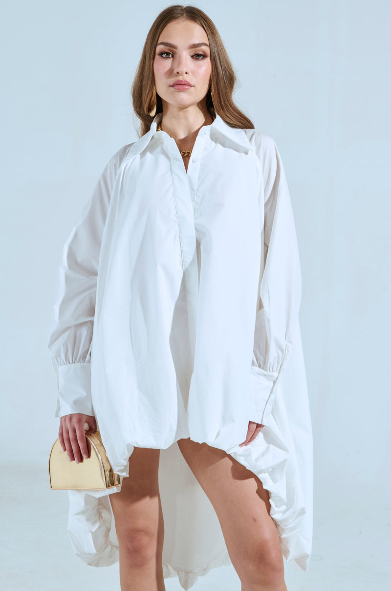 A person stands confidently in the SHE MEANS BUSINESS MINI DRESS, a flowing white shirt dress with an asymmetrical hem made from a cotton blend, holding a small beige clutch. They have long hair and a neutral expression set against a blue background.
