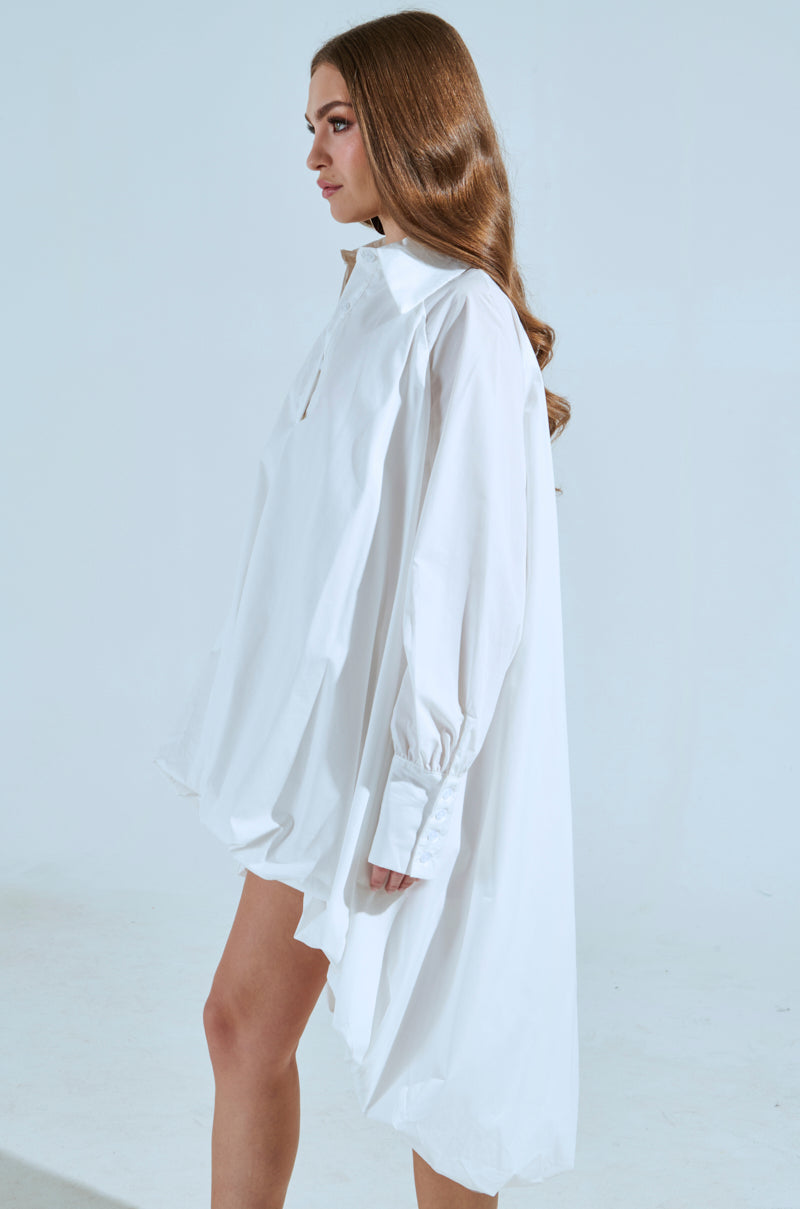 A woman with long brown hair stands in profile against a plain light blue-grey background, wearing the SHE MEANS BUSINESS MINI DRESS—a loose-fitting white button-down mini dress featuring long sleeves and a turned-up collar.