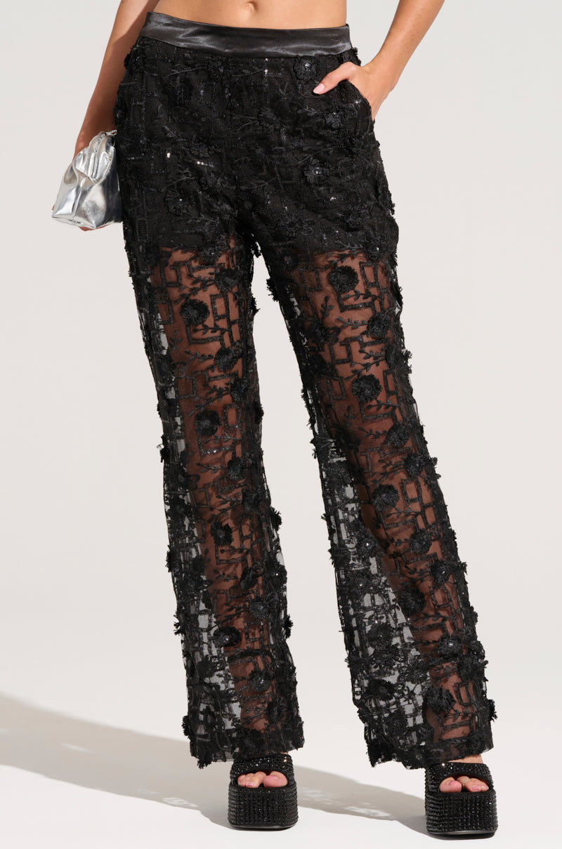 A person is modeling the AKIRA Label Celebration Time Lace Pants in black, featuring a high-rise design with sheer sequin-embellished fabric and a solid waistband. They are holding a small silver clutch in one hand and wearing platform shoes. The background is plain and light-colored.