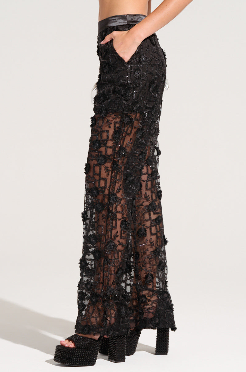 A model is wearing the CELEBRATION TIME LACE PANTS IN BLACK, which are black sequined, sheer, wide-leg pants featuring intricate embroidery and a high-rise design. Their left hand rests in a pocket as they stand in profile, paired with platform shoes adorned with stud detailing.
