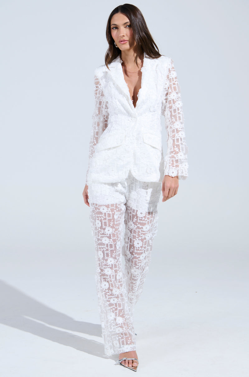 A woman models a sheer white lace pantsuit with a deep V-neck and long sleeves, similar to the CELEBRATION TIME BLAZER, showcasing intricate patterns. Her look is completed with silver strappy heels on a plain light gray background.