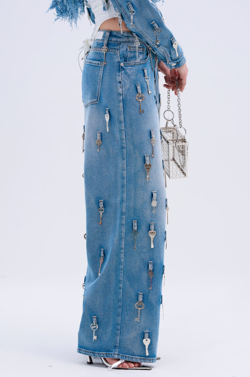 Someone wears AKIRA Label Key To The City Relaxed Fit Jeans adorned with small metal keys, a matching denim jacket, and white heeled sandals, holding a metallic handbag. The jeans have a mid-rise fit against a plain white background.