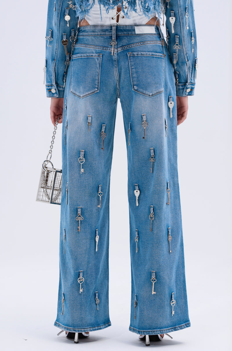 Someone is seen from the back wearing KEY TO THE CITY RELAXED FIT JEANS, featuring wide blue legs adorned with silver keys. They hold a small metallic purse against a plain, light-colored background.