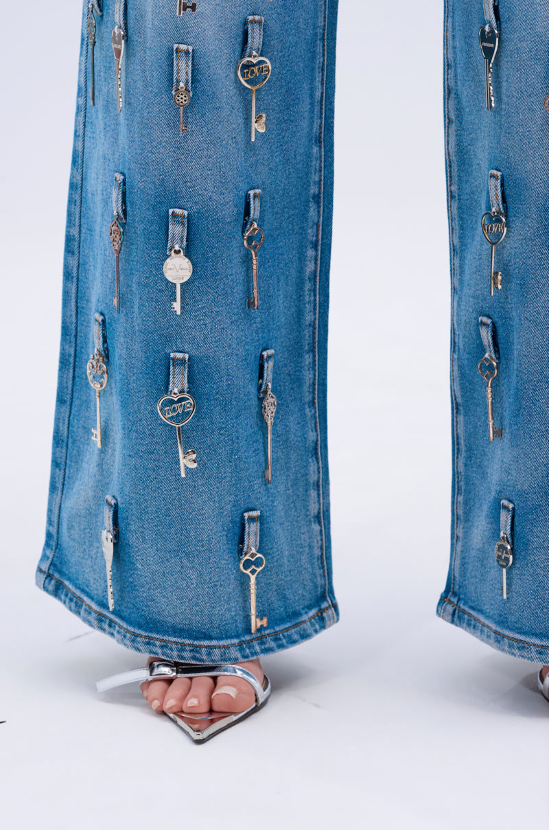 Close-up of KEY TO THE CITY RELAXED FIT JEANS in blue denim, detailed with multiple metallic keys along the legs; styled with silver high-heeled square-toe sandals, photographed against a plain white background.