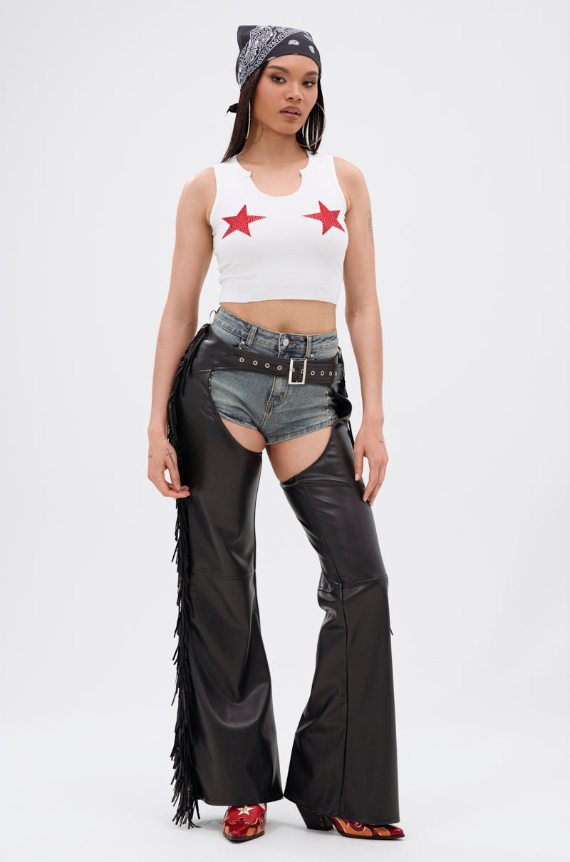 A woman poses in a white crop top, red star details, denim shorts, and RODEO FRINGE FAUX LEATHER CHAPS—black faux leather with fringe—plus red shoes for a bold Western-chic look against a plain white background.