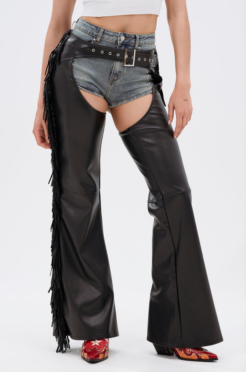 A person wears a white crop top, denim shorts, and RODEO FRINGE FAUX LEATHER CHAPS with one fringed leg for a bold Western look. Styled with a black belt and red patterned boots. Image is cropped at the shoulders.