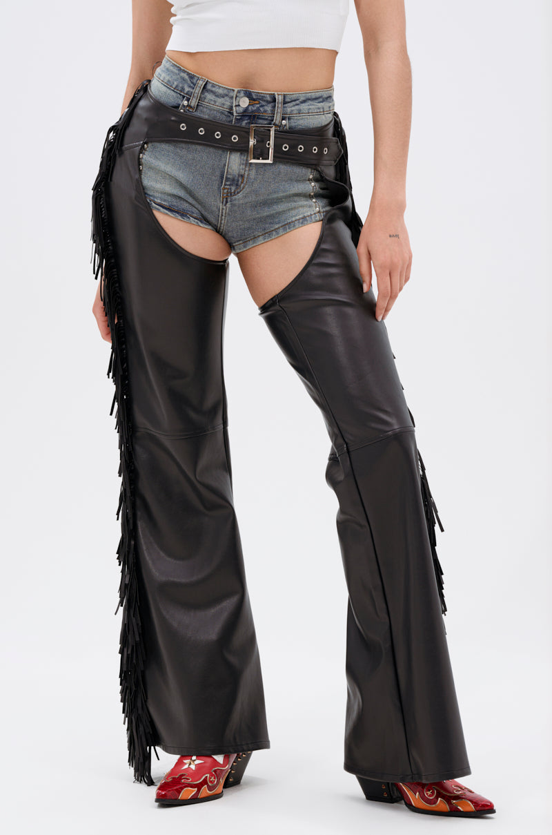 Model wears white crop top, denim shorts, and RODEO FRINGE FAUX LEATHER CHAPS styled with a studded belt and red flame cowboy boots for a bold Western-chic look against a plain background.