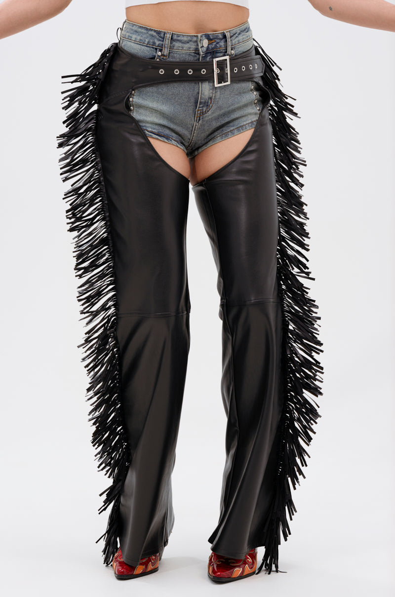 Someone models denim shorts with a black belt under RODEO FRINGE FAUX LEATHER CHAPS, which have long side fringe. Red patterned heels finish the look, all shown against a plain white background.