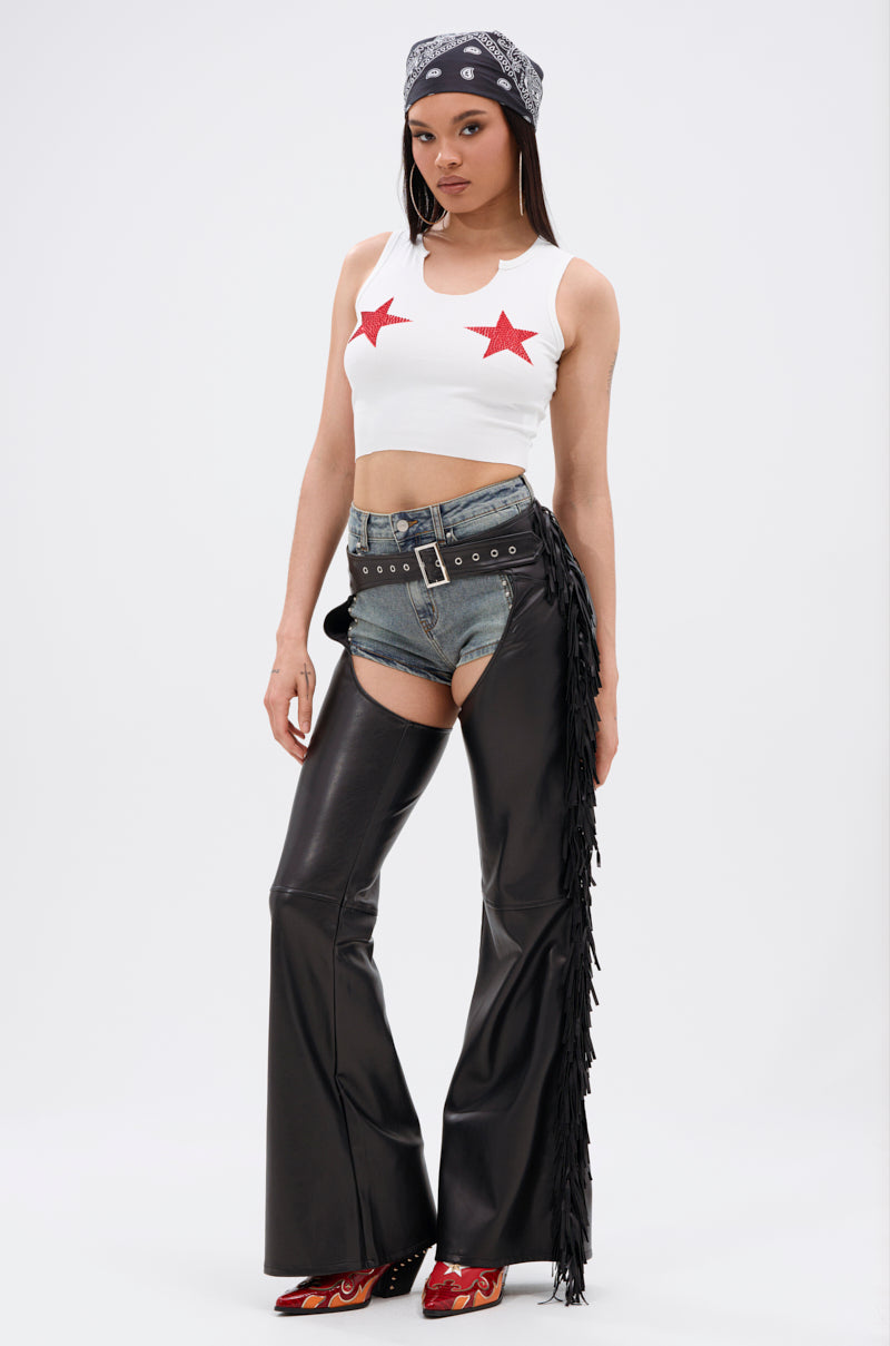 A woman stands confidently in a white crop top with red stars, denim shorts, Western-chic bottoms, RODEO FRINGE FAUX LEATHER CHAPS over her shorts, a double belt, red cowboy boots, and a black bandana on her head.