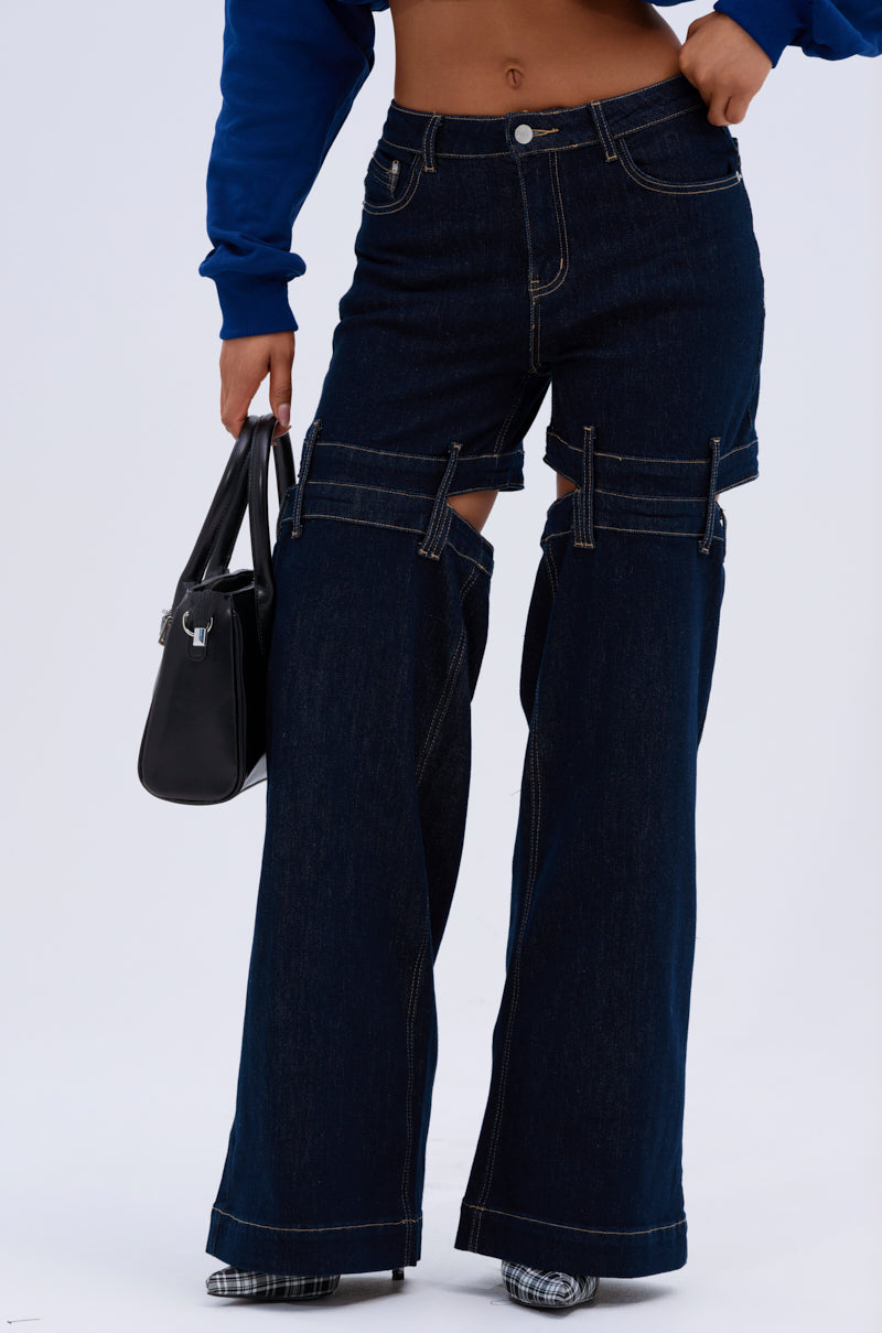 A person wearing the LOCKED UP DENIM PANT—dark blue wide-leg jeans with a mid-rise fit and detachable upper thigh sections—holds a black handbag and stands against a light background.