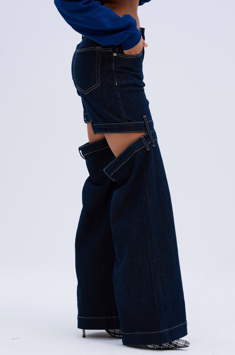 A person styles the LOCKED UP DENIM PANT, featuring dark-wash wide legs and bold thigh and knee cutouts, with a blue top and checked pointed heels. The mid-rise fit pops against the plain light gray background.