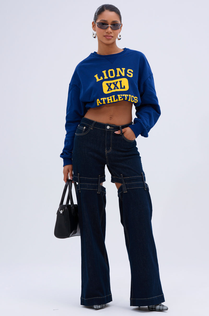 A woman poses confidently in sunglasses, a blue "LIONS XXL ATHLETICS" cropped sweatshirt, LOCKED UP DENIM PANT with wide-leg silhouette and zipper details, checkered shoes, and a black handbag against a plain background.