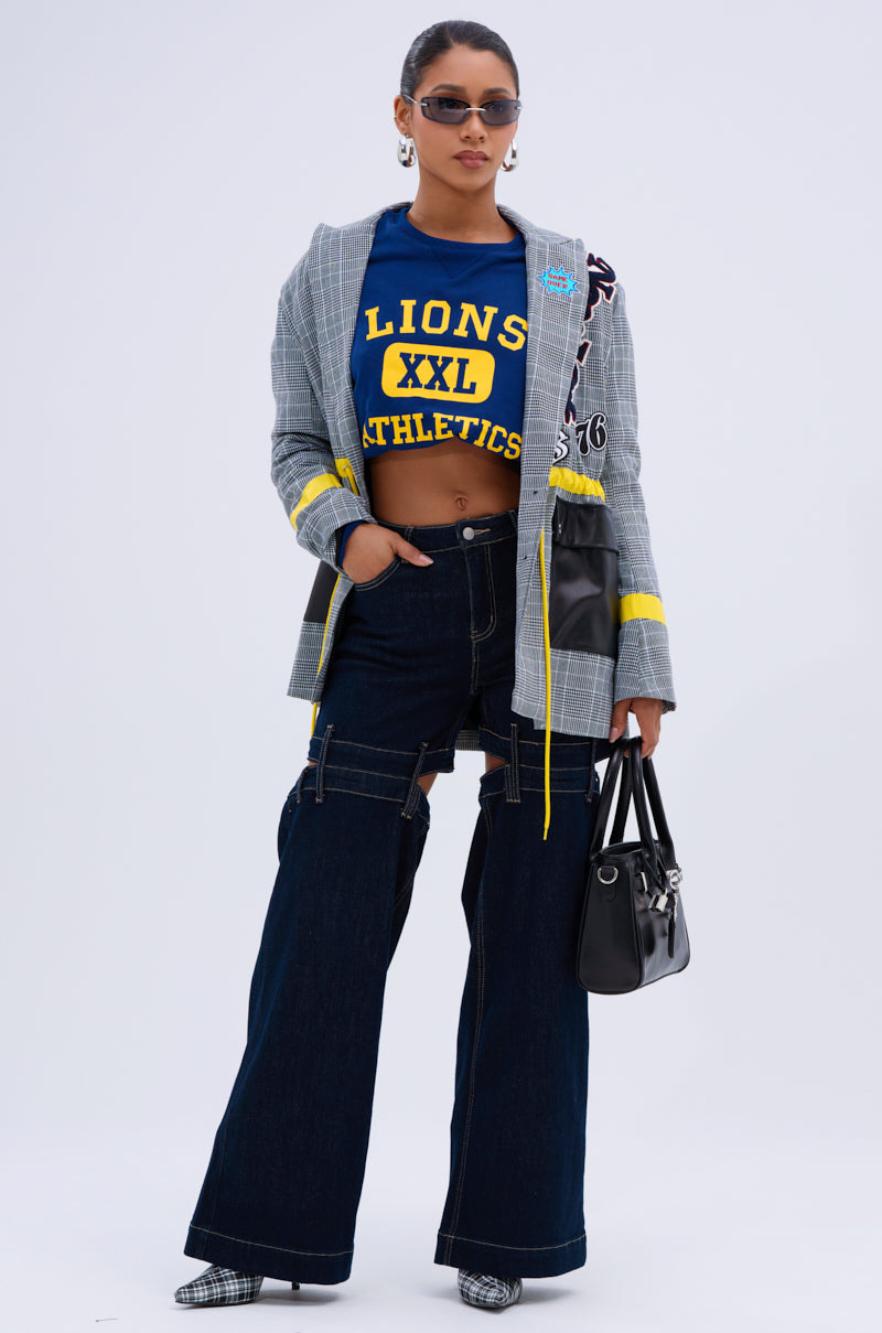 A woman stands confidently wearing sunglasses, a blue "Lions XXL Athletics" crop top, a plaid blazer, checkered shoes, and holds a black handbag. Her look features the LOCKED UP DENIM PANT in a wide-leg silhouette with cutouts against a plain background.