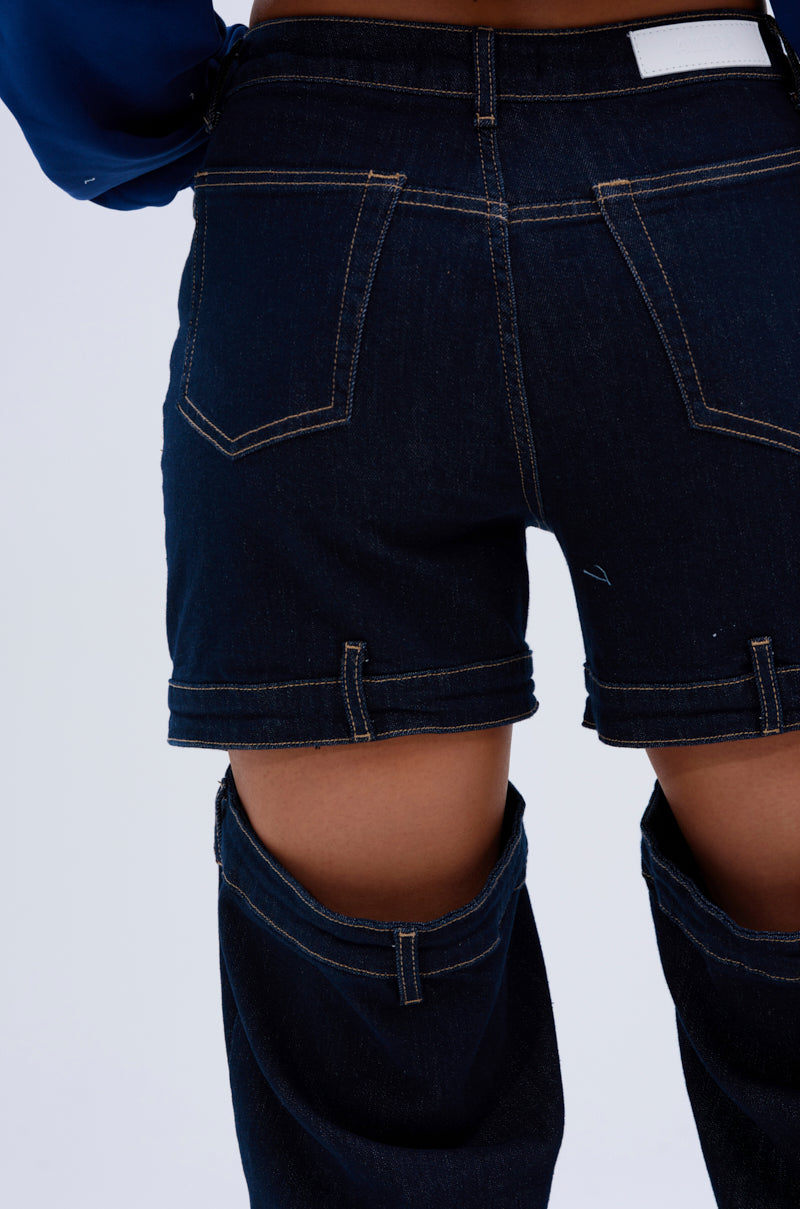 Wearing the LOCKED UP DENIM PANT—dark, mid-rise denim with detachable legs unzipped into shorts—this photo captures a back view from waist to knees, emphasizing the unique wide-leg silhouette and exposed thighs.