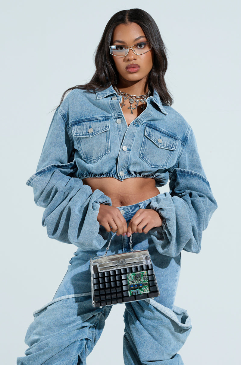 A woman exudes confidence in a STACKED SLEEVE DENIM JACKET and matching pants, accessorized with a small textured handbag. Her long hair flows freely, complemented by glasses and layered necklaces for a chic ensemble.