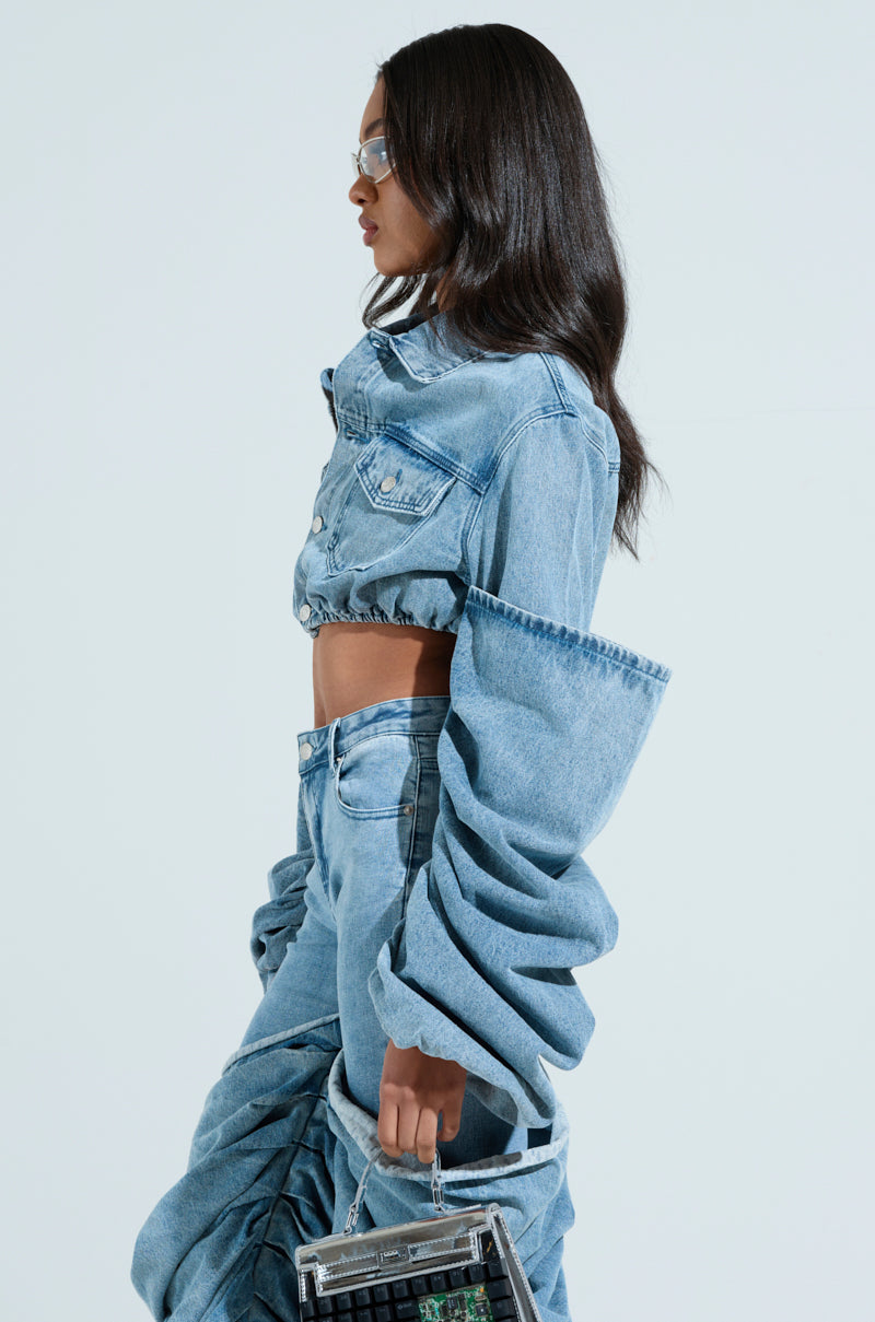 A woman with long dark hair is seen in profile against a plain background. She wears a denim crop top, oversized denim pants, and the Stacked Sleeve Denim Jacket. She's accessorized with sunglasses and holds a small transparent handbag.