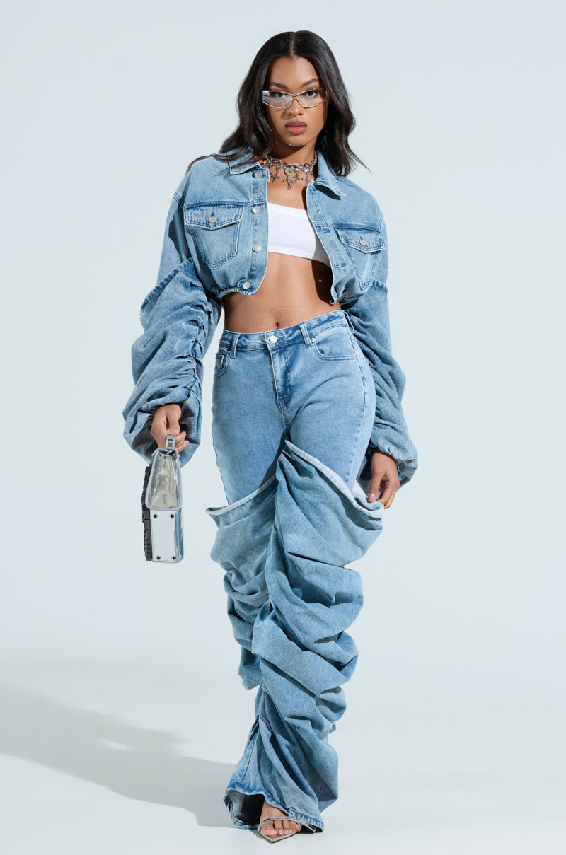 A woman wears a STACKED SLEEVE DENIM JACKET in a cropped, oversized fit with ruched denim pants and a white crop top. She accessorizes with glasses, strappy heels, a small silver handbag, and a gold necklace against a plain, light backdrop.