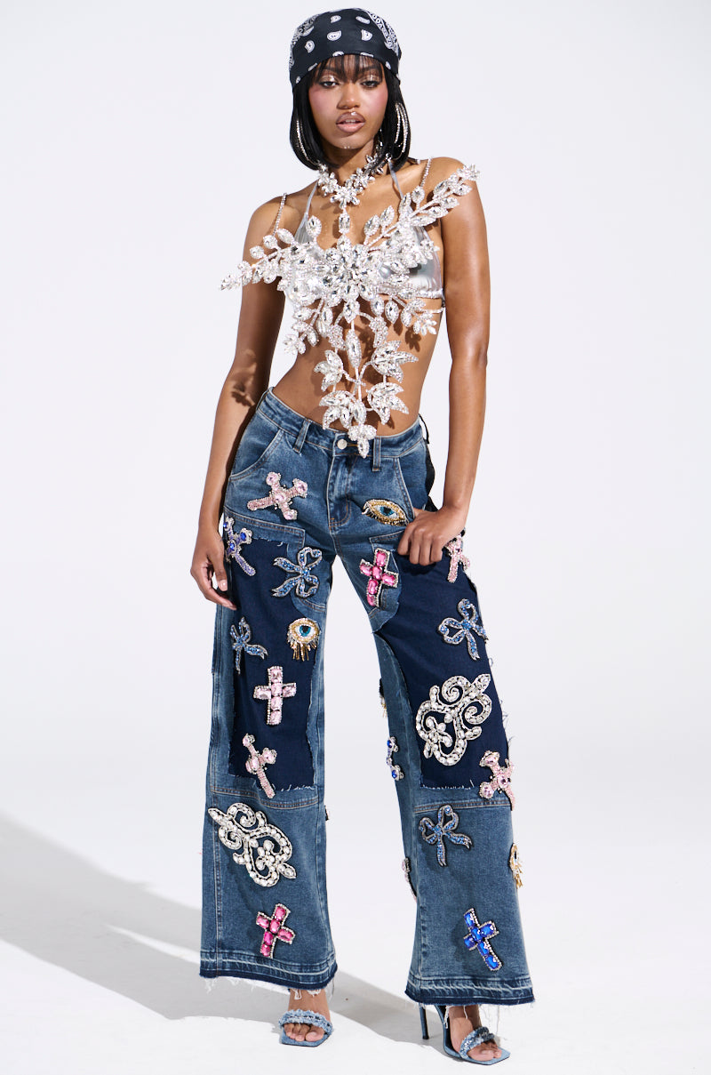 Confident in their unique outfit, a person showcases the WILD AND FREE RHINESTONE DENIM—rhinestone-studded wide-leg pants with colorful patches—and pairs it with a decorative, sheer top, patterned head scarf, and chic heels against a minimalistic light background.