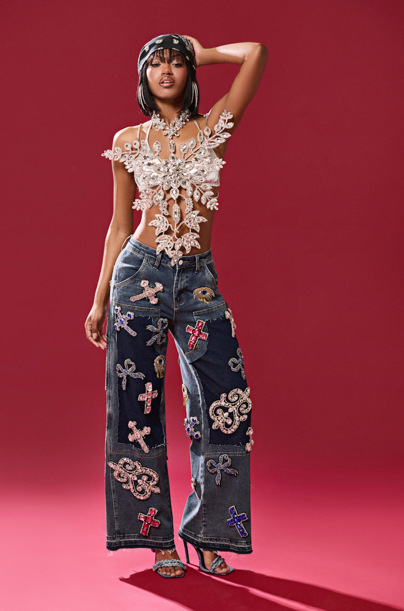 In front of a red background, a person models WILD AND FREE RHINESTONE DENIM adorned with colorful appliqués and paired with a decorative floral top. Their pose is accentuated by a headband and high heels, one hand resting on their head.