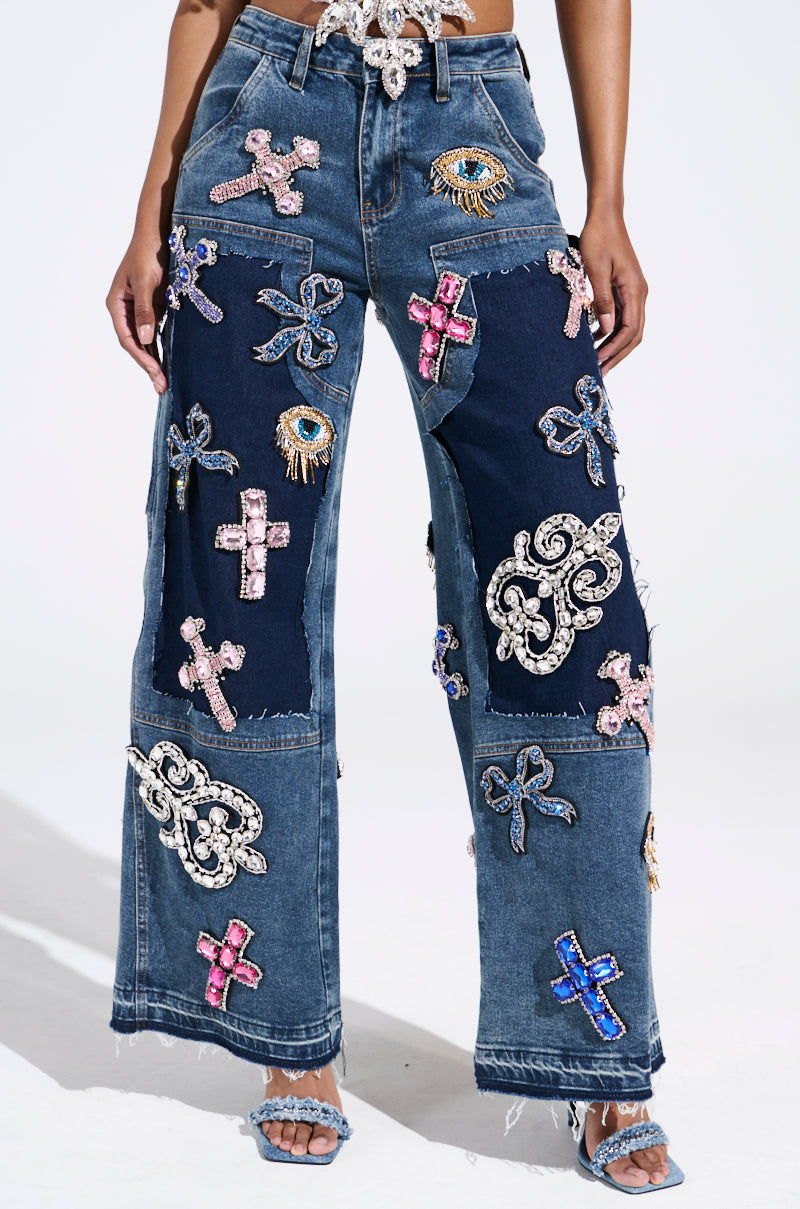 Sporting the WILD AND FREE RHINESTONE DENIM, an individual showcases wide-leg jeans adorned with colorful embroidered patches like crosses and eyes, set against dark and light denim. Shimmering rhinestones pair brilliantly with embellished blue sandals.