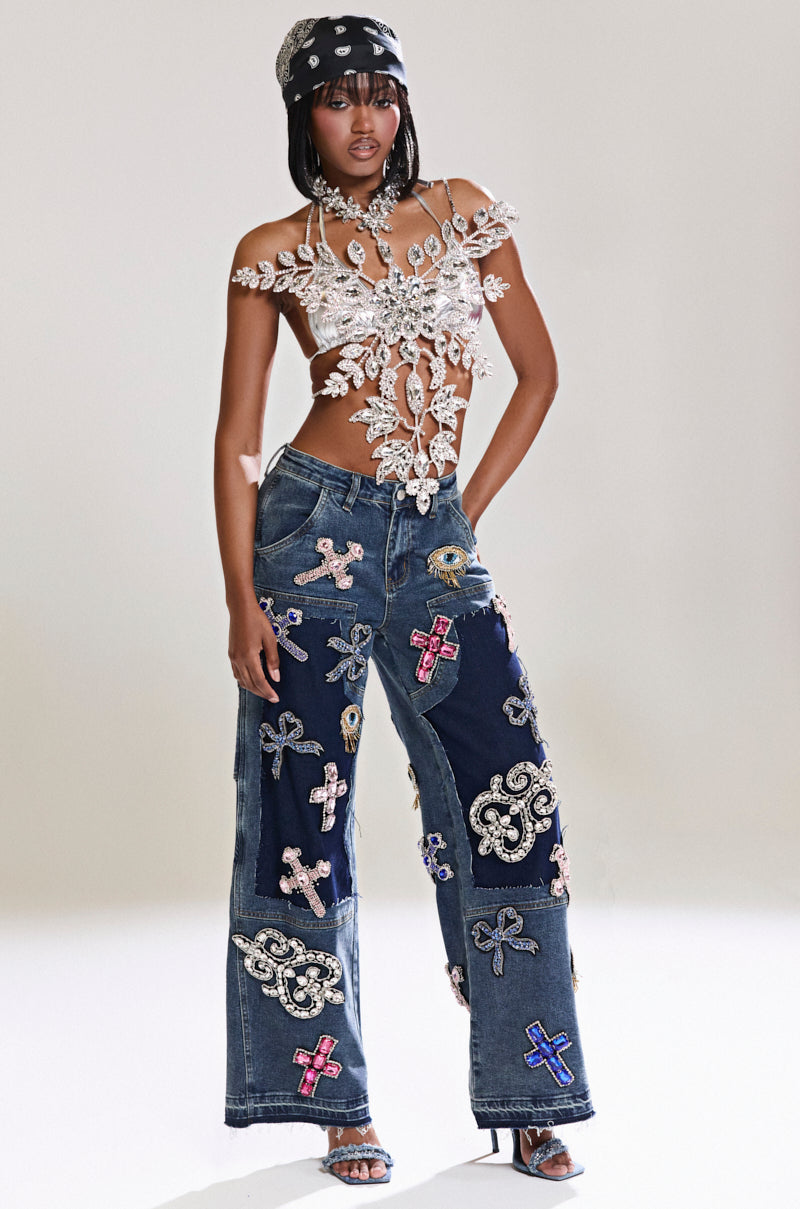 A person confidently poses in an outfit featuring the WILD AND FREE RHINESTONE DENIM, pairing a unique white lace top with wide-leg jeans adorned with colorful embroidered patches, complemented by a black bandana and heels against a neutral background.