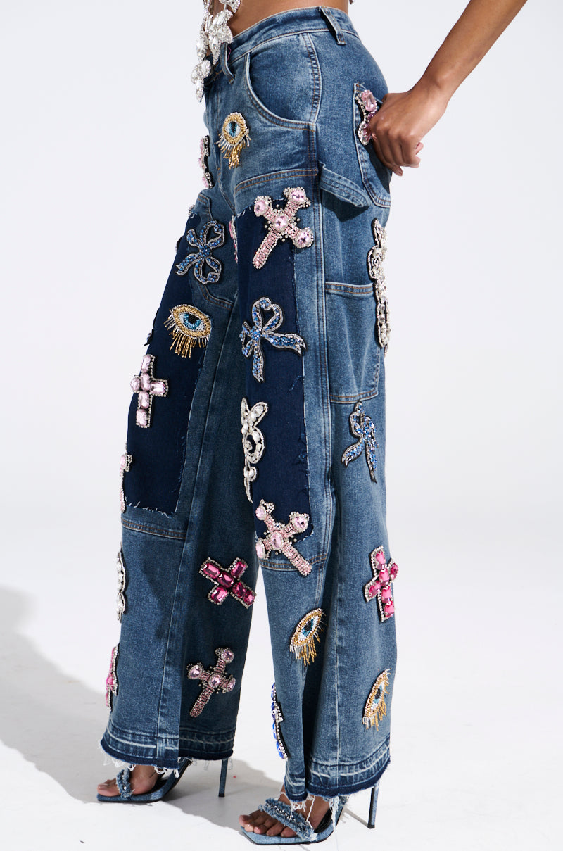 A person showcases the WILD AND FREE RHINESTONE DENIM, featuring colorful crosses, eyes, and rhinestone accents on a patchwork wide-leg silhouette. Styled with high-heeled sandals against a plain backdrop, the vibrant and intricate designs are highlighted.
