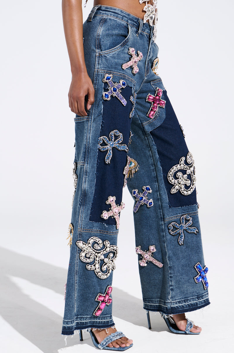 Someone is wearing the WILD AND FREE RHINESTONE DENIM jeans, featuring colorful sequin cross patches in various sizes on dark blue panels. The wide-leg silhouette pairs seamlessly with blue heeled sandals.