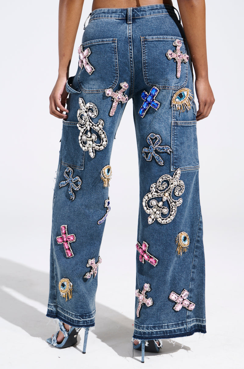 A person wearing WILD AND FREE RHINESTONE DENIM, embellished with colorful sequin patches of crosses, swirls, and eyes, is seen from the back. They stand poised in high-heeled shoes against a plain white backdrop.