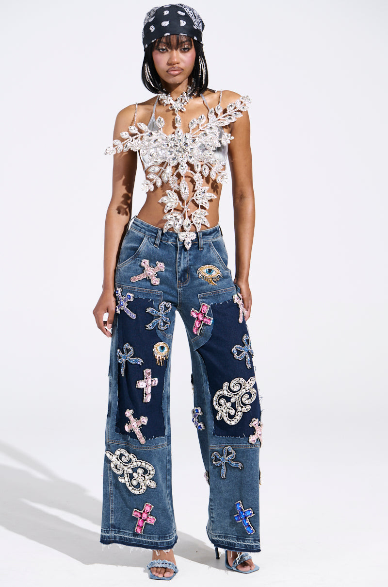 A person poses in a fashion-forward outfit featuring a delicate top and WILD AND FREE RHINESTONE DENIM wide-leg jeans, adorned with embroidered patches. They accessorize with a bandana and heels, standing against a plain background.