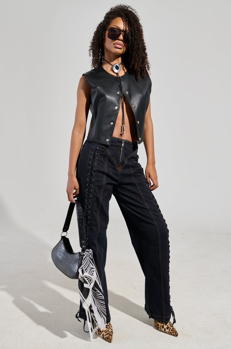 Dressed confidently, the individual wears sunglasses with curly hair and a striking outfit that includes a black leather vest and COULD OF BEEN ME DENIM PANT—high-waisted dark jeans featuring eye-catching lace-up paneling. Completing the look is a black handbag and animal-print shoes, all set against a minimal and neutral background.
