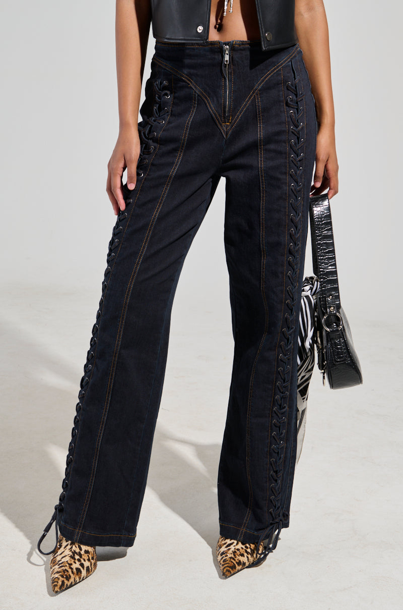 A person wearing the "Could of Been Me Denim Pant", which features a black lace-up design and a straight leg silhouette, paired with a black sleeveless top and leopard-print heels. They clutch a black textured handbag and hold a zebra-patterned piece against the light-colored background.