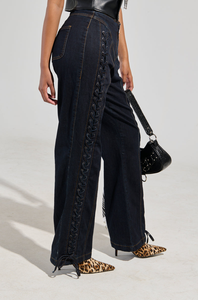 A person wearing the COULD OF BEEN ME DENIM PANT, featuring high-waisted black jeans with lace-up paneling and a straight leg silhouette, paired with a black top, leopard print ankle boots, and holding a black handbag. The jeans elegantly extend to the ground.
