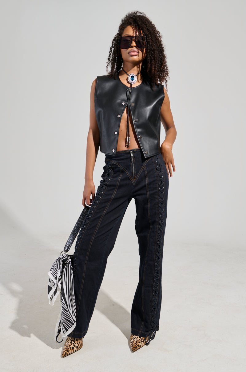 A person with curly hair wearing sunglasses poses confidently. They are dressed in a sleeveless black leather vest and COULD OF BEEN ME DENIM PANT, which features lace-up paneling. They hold a bag with a zebra print scarf and are wearing leopard-print shoes.