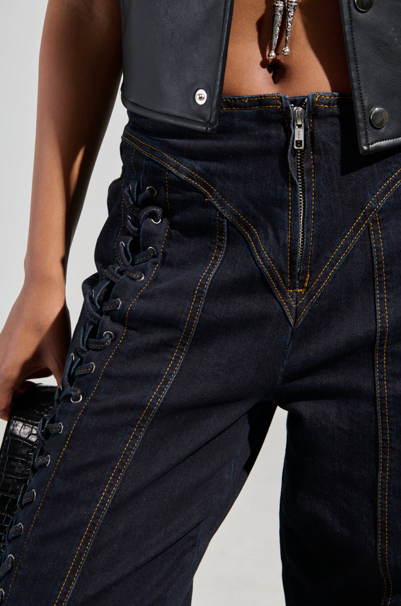 Close-up of a person wearing the "COULD OF BEEN ME DENIM PANT," which are high-waisted dark denim jeans with prominent yellow stitching and lace-up paneling on the sides. The individual pairs them with a black leather crop top and holds a textured black handbag.