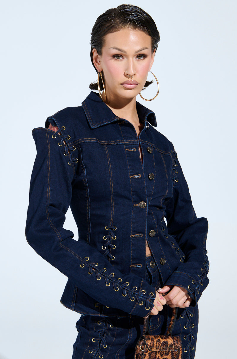 A person confidently wearing the THATS ON ME BUTTON DOWN TOP, a dark denim piece with lace-up detailing on the sleeves and sides, stands against a light background. They have hoop earrings and styled short hair, looking directly at the camera.