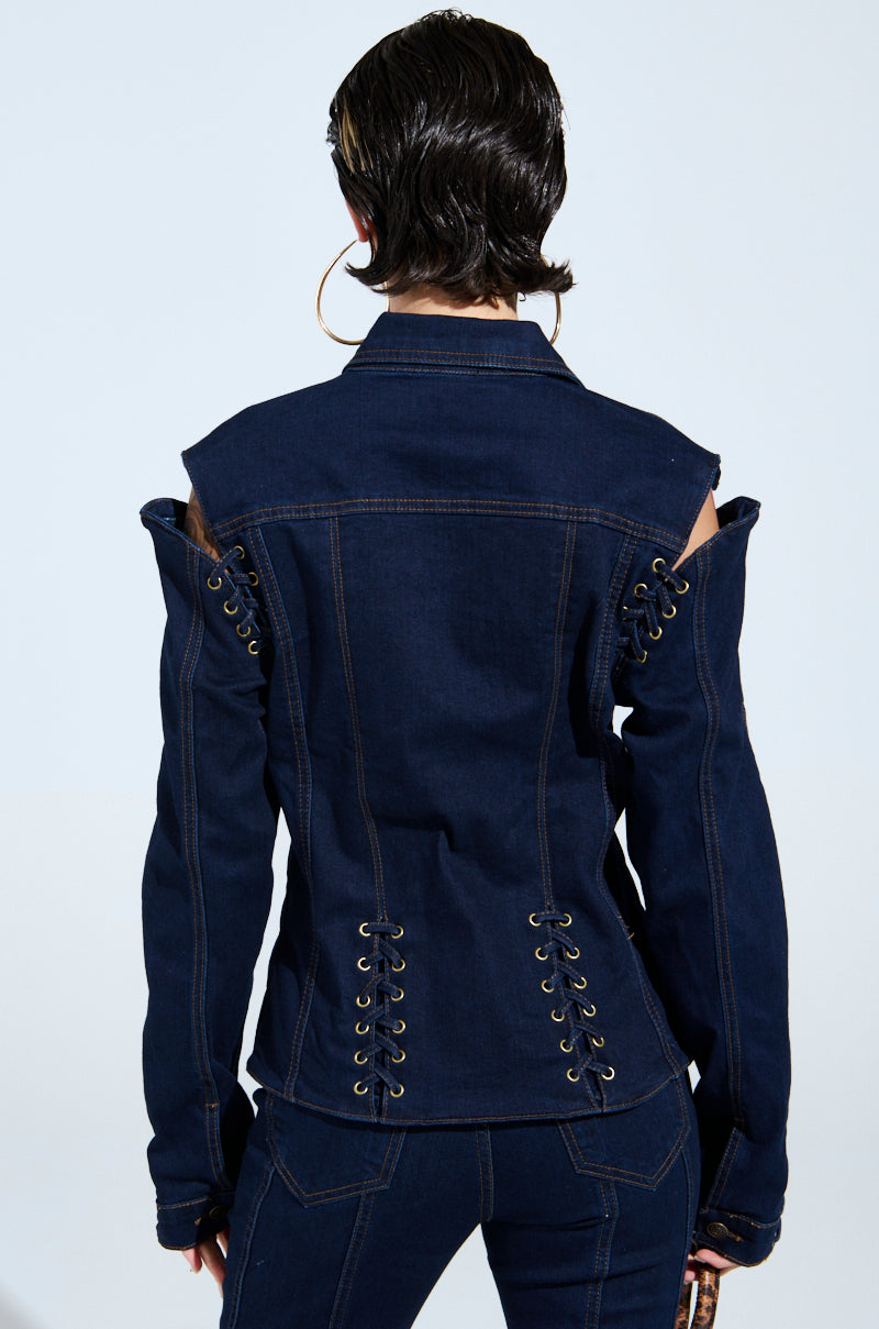 A person with short hair is wearing the THATS ON ME BUTTON DOWN TOP, a dark denim jacket featuring chic lace-up detailing along the sleeves and back, with modern shoulder cutouts, perfectly complemented by matching denim pants.