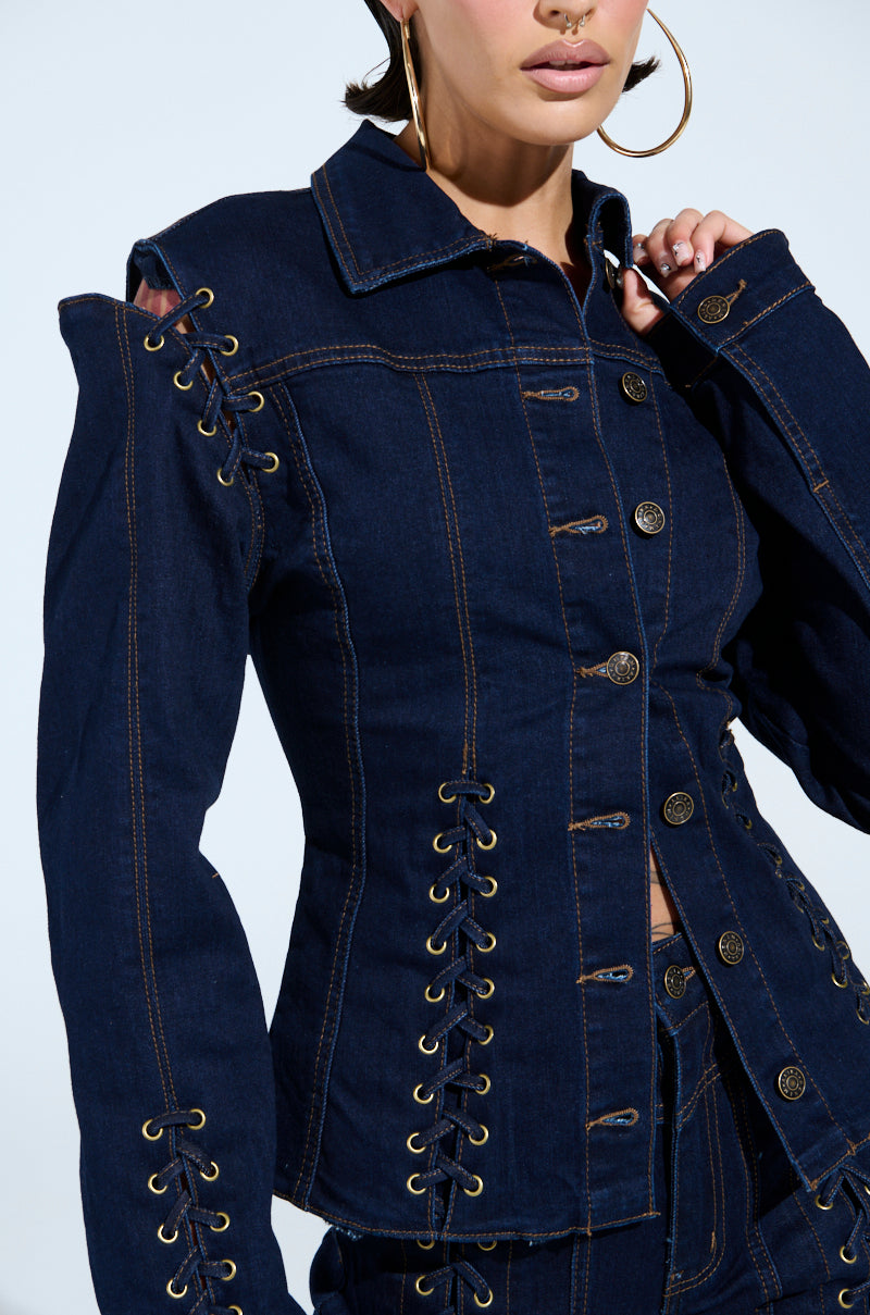 The image features a person modeling the chic "THATS ON ME BUTTON DOWN TOP," a dark denim jacket with intricate lace-up detailing and modern metal buttons. Trendy shoulder cut-outs complement the look, accessorized with large hoop earrings against a light backdrop.
