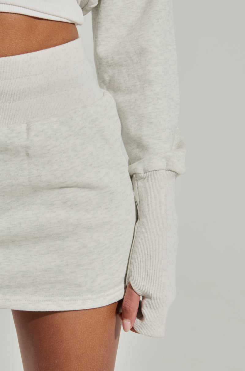A close-up of a person wearing the STINGY WITH MY TIME MINI SKIRT, a matching high-rise mini skirt and light gray long-sleeve crop top made from a stretchy cotton blend. The person's hand is partially visible, with the long sleeve extending over part of the hand. The background is plain and light-colored.